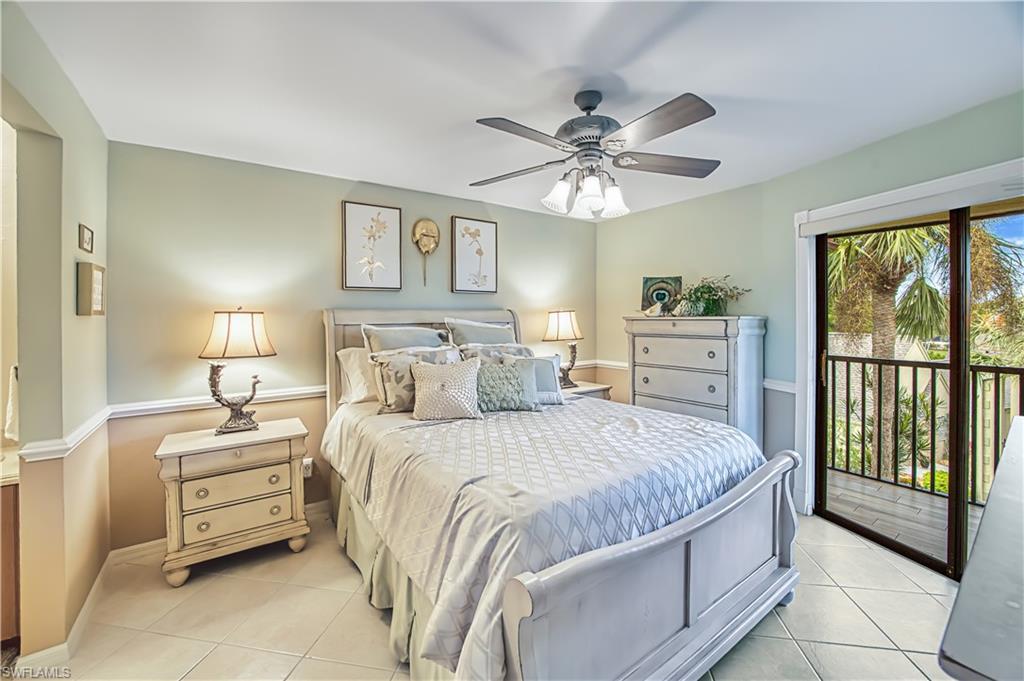 Master bedroom featuring a ceiling fan with integrated lighting, dual nightstands with wood-finish surfaces, neutral wall paint in soft earth tones, tile flooring throughout, and French doors opening to a covered balcony with tropical landscaping views