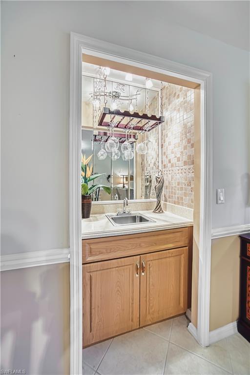 Recessed wet bar featuring light wood cabinetry, a stainless steel sink, tiled backsplash, and an overhead wine glass rack
