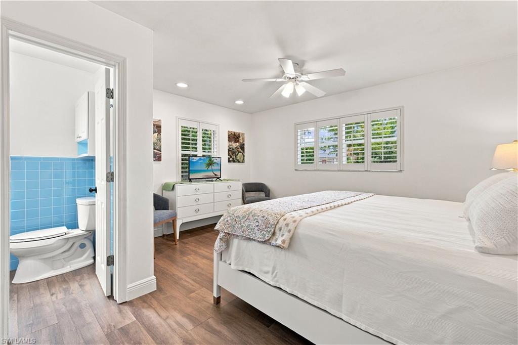 Wood-finish flooring throughout, white walls, ceiling fan, window with white plantation shutters, and an adjacent bathroom with blue tiled wainscoting