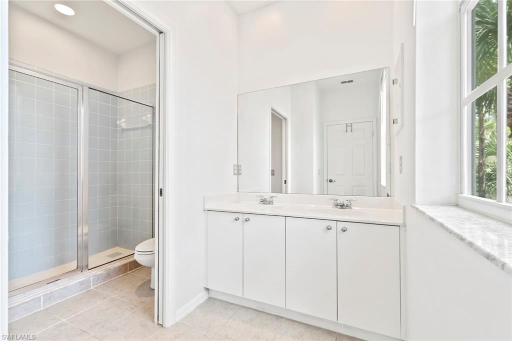 Dual vanity with white cabinetry and chrome hardware, a large mirror, a window featuring multiple panes, and a glass-enclosed shower with tiled walls