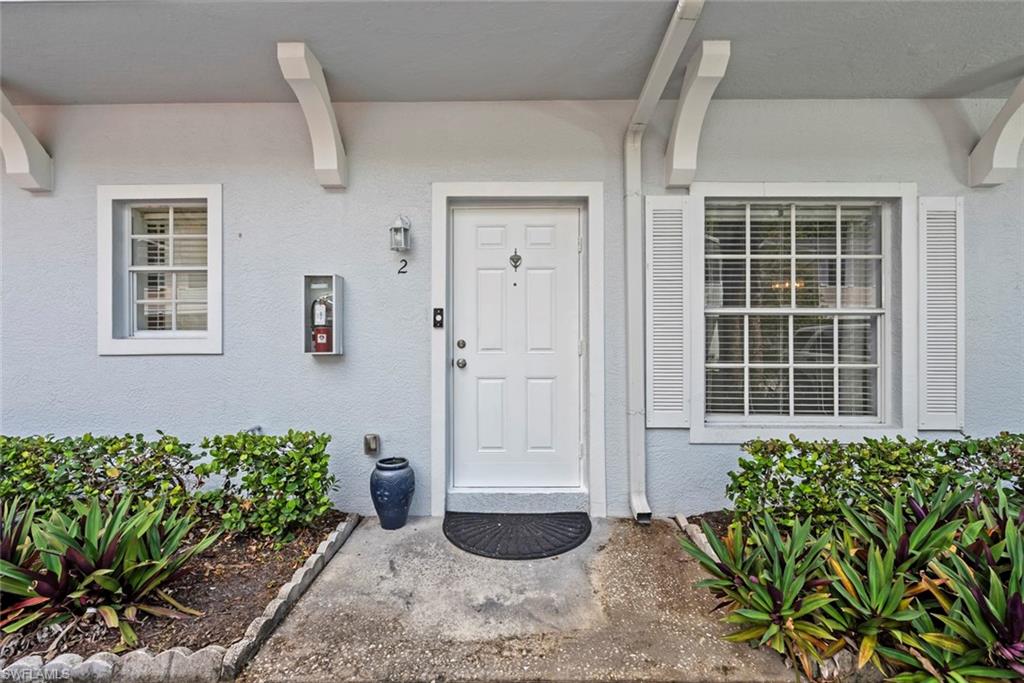 White paneled entry door, exterior wall lantern, decorative window shutters, foundation planting, and concrete walkway