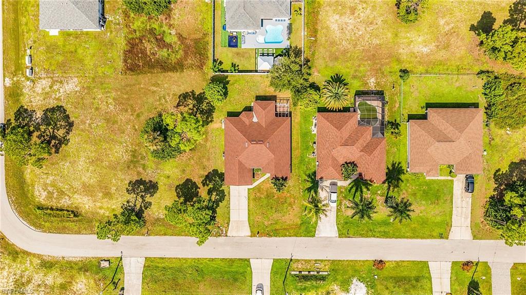 Aerial perspective of a residential street with multiple properties, featuring a corner lot with undeveloped land, houses with brown roofs, and a property with a visible swimming pool