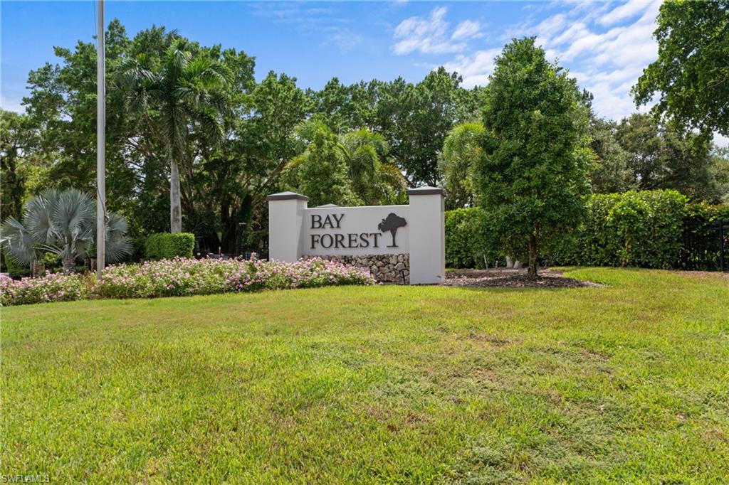Community entrance sign featuring a stone base and "BAY FOREST" lettering, surrounded by lush landscaping with flowering shrubs, mature trees, and a manicured lawn
