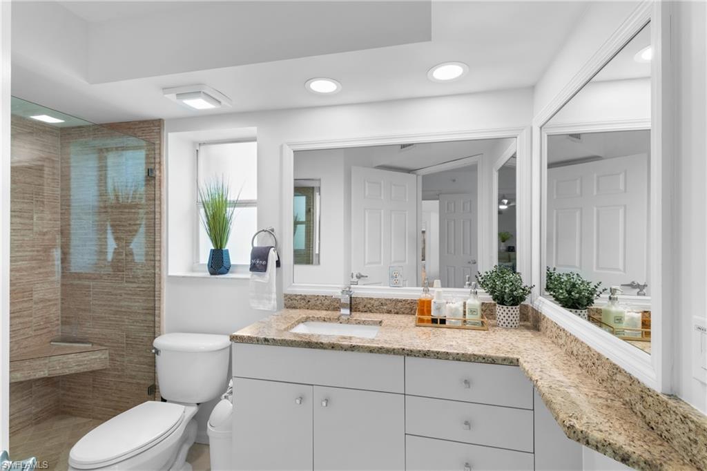 Glass-enclosed shower with a tiled bench, white vanity with a single sink, light-toned stone countertop, and two large wall-mounted mirrors