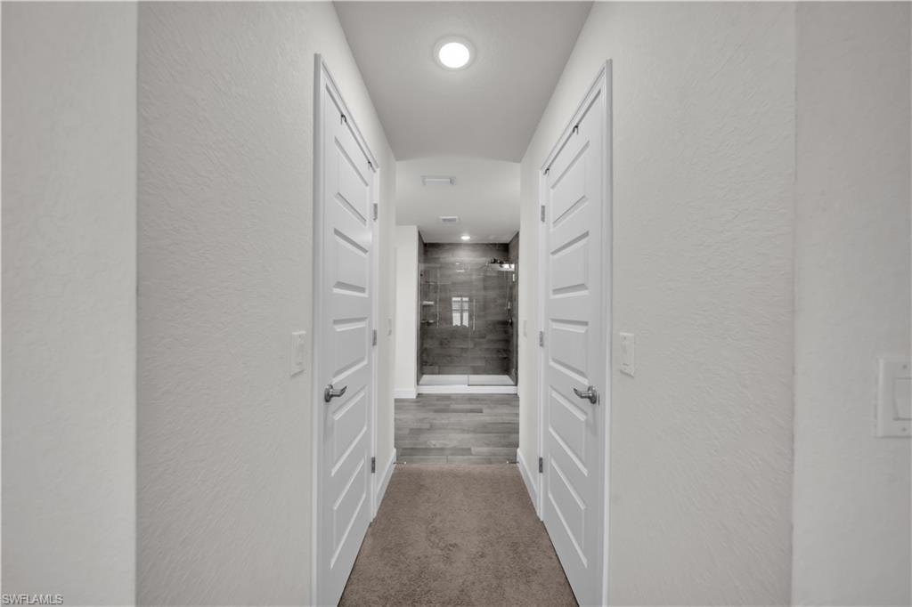 Recessed lighting illuminates a hallway with white paneled doors and textured walls, leading to a bathroom with a glass-enclosed shower and wood-finish flooring