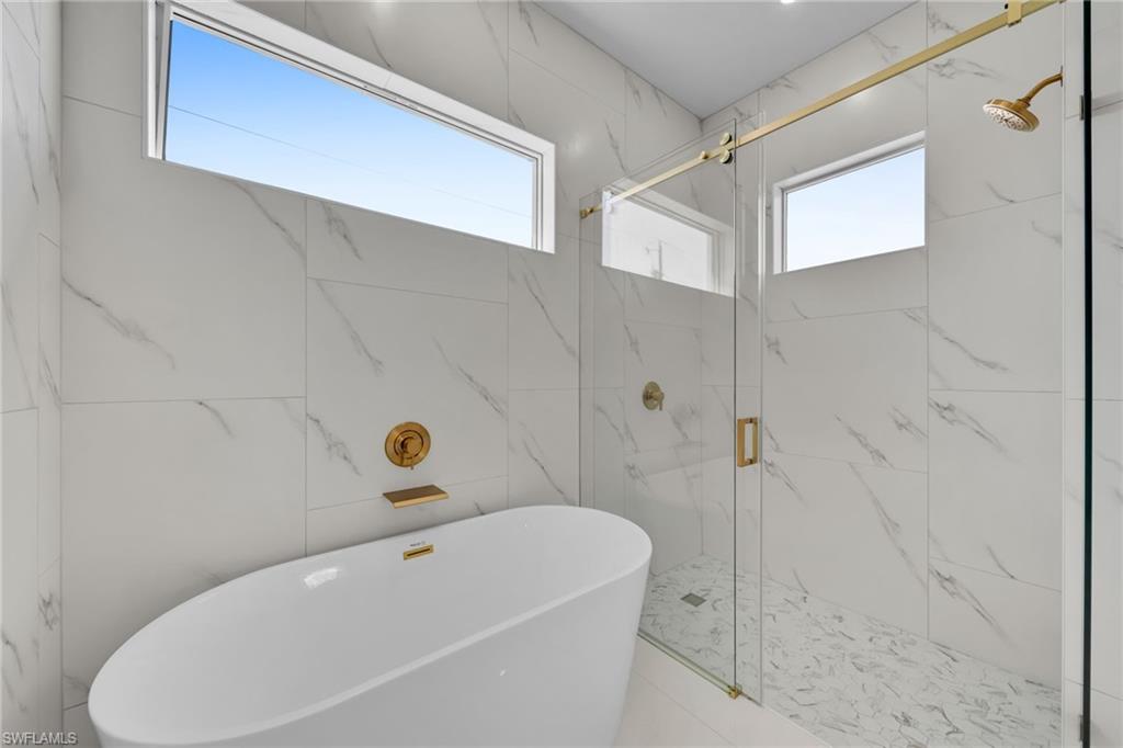 Freestanding soaking tub with a wall-mounted faucet, a separate glass-enclosed shower with gold-tone hardware, and large format wall tiling