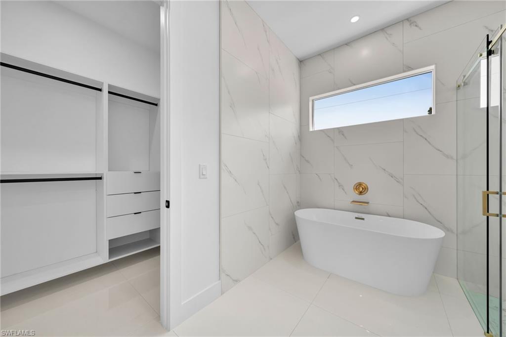 Freestanding soaking tub with gold-finish fixtures, a walk-in shower with glass enclosure, large format tile flooring, and a built-in closet system