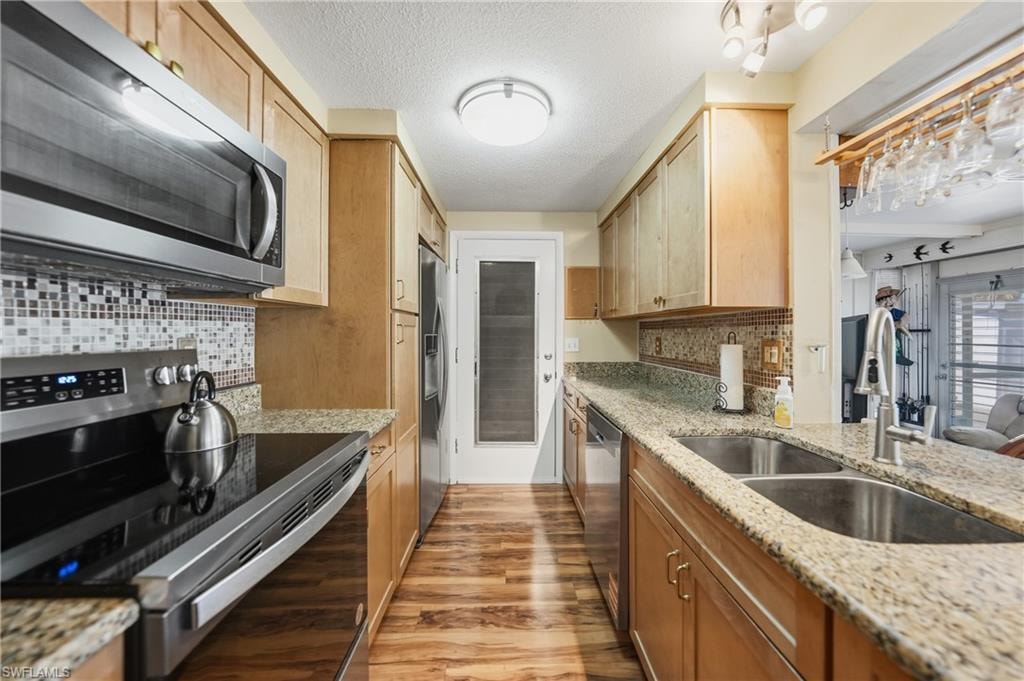 Galley kitchen featuring light wood cabinetry, stone countertops, stainless steel appliances, a mosaic tile backsplash, and wood-finish laminate flooring