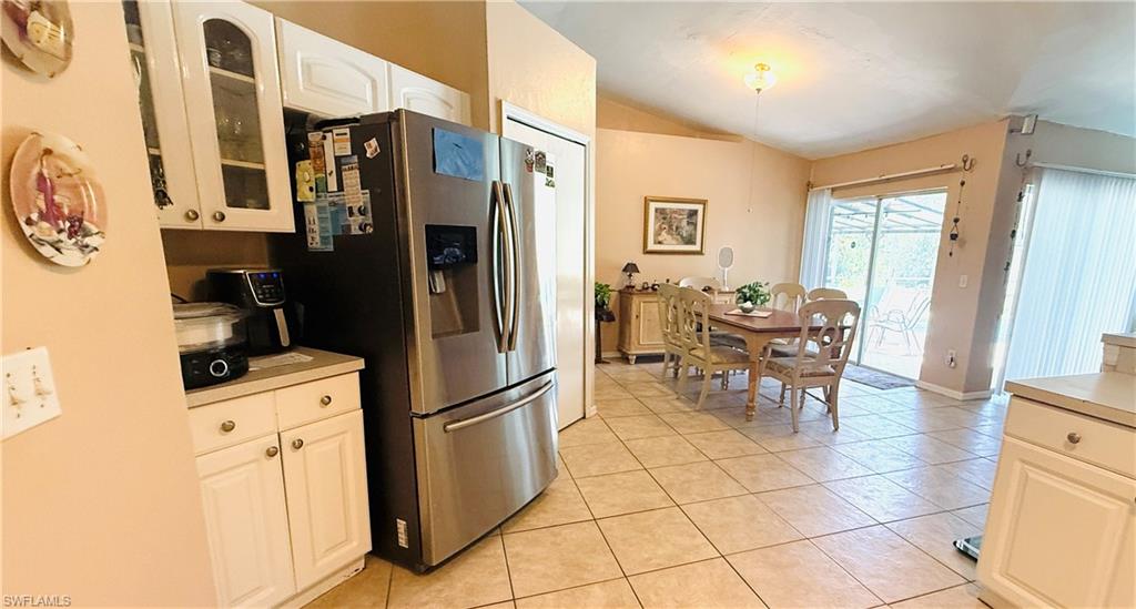 Open kitchen and dining area featuring stainless steel refrigerator with French doors, white cabinetry with glass-front upper cabinets, tile flooring throughout, and abundant natural light from multiple windows and glass doors leading to the adjoining space