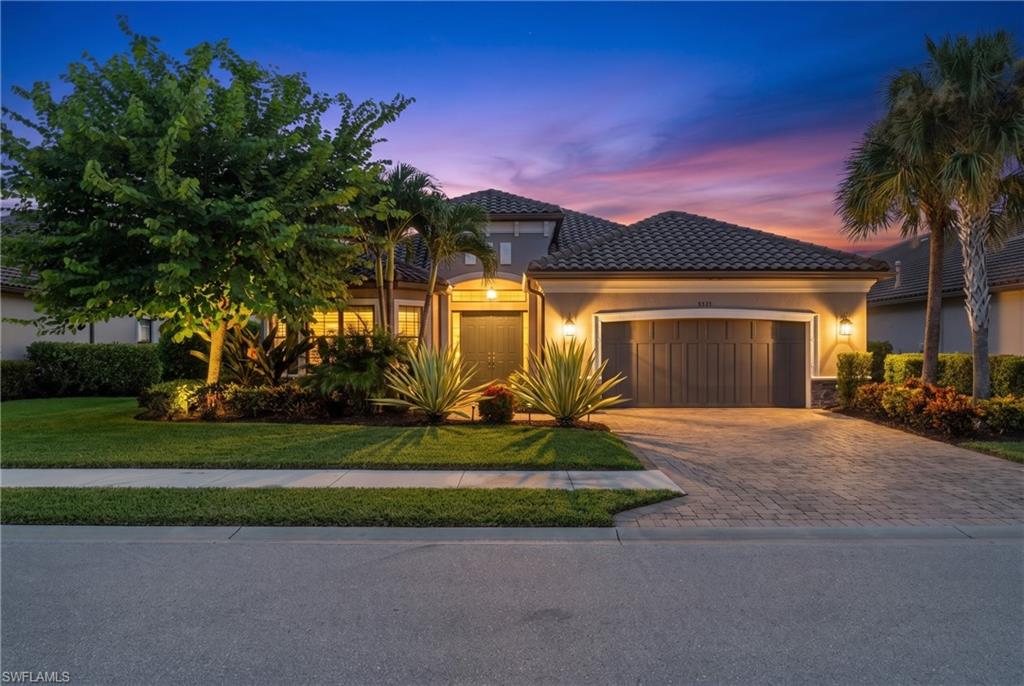 Mediterranean-style architecture with a tile roof, an integrated two-car garage, paver driveway, and landscaped front yard with mature trees