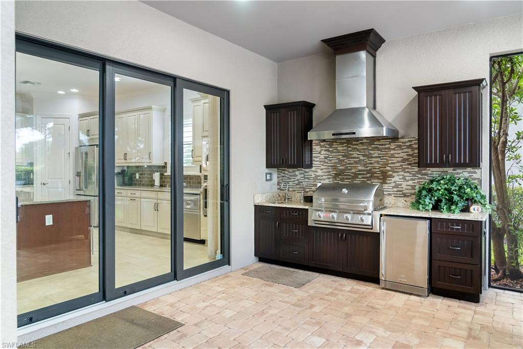 Outdoor kitchen featuring built-in grill, stainless steel range hood, stone tile backsplash, wood-finish cabinetry, and light-toned countertop