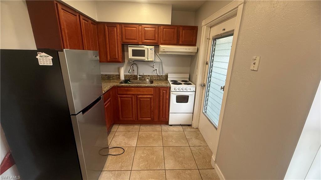 Kitchen with white appliances, light tile patterned floors, under cabinet range hood, brown cabinetry, and dark stone countertops