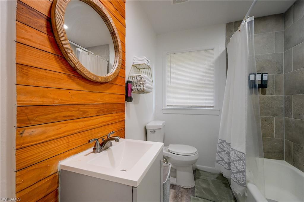 Vanity with an integrated sink and brushed nickel faucet, wood-paneled accent wall, round mirror, tiled shower surround, and a window with blinds