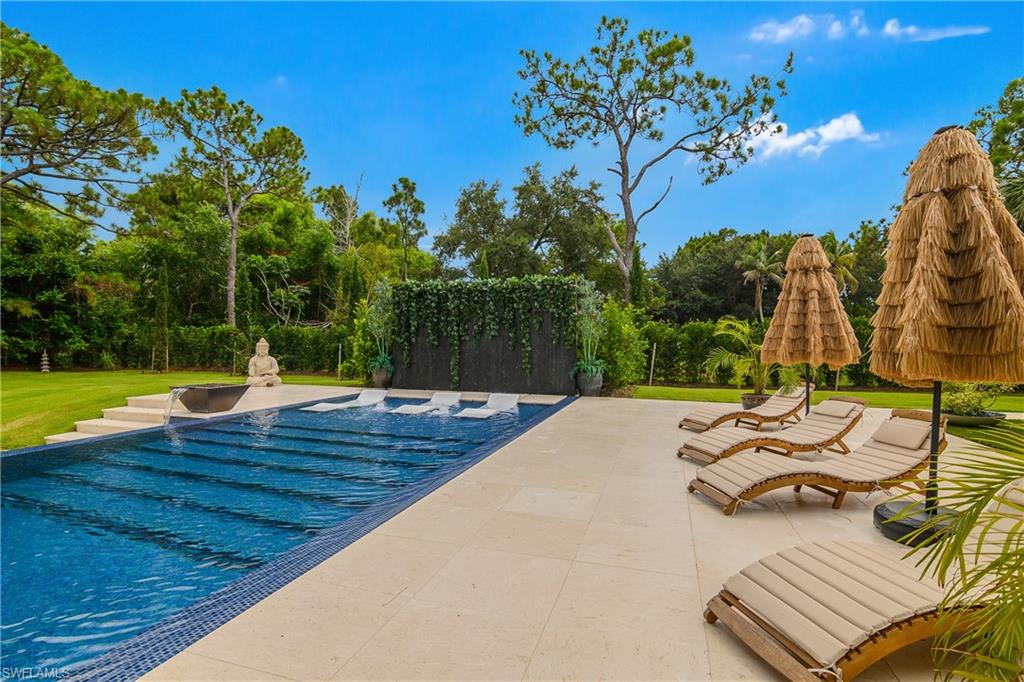 Expansive pool deck with light-toned pavers, a dark-tiled pool with integrated steps, and a water feature with an attached planter