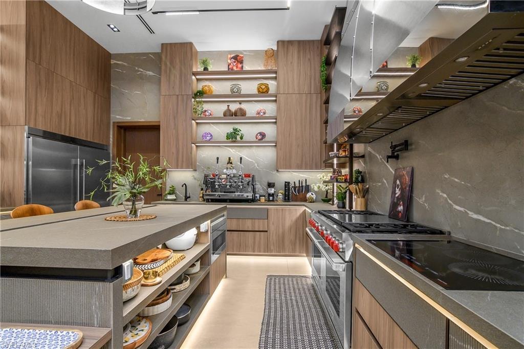Gourmet kitchen featuring wood-finish cabinetry, stainless steel appliances, a built-in range hood, a large island with integrated shelving, and stone countertops