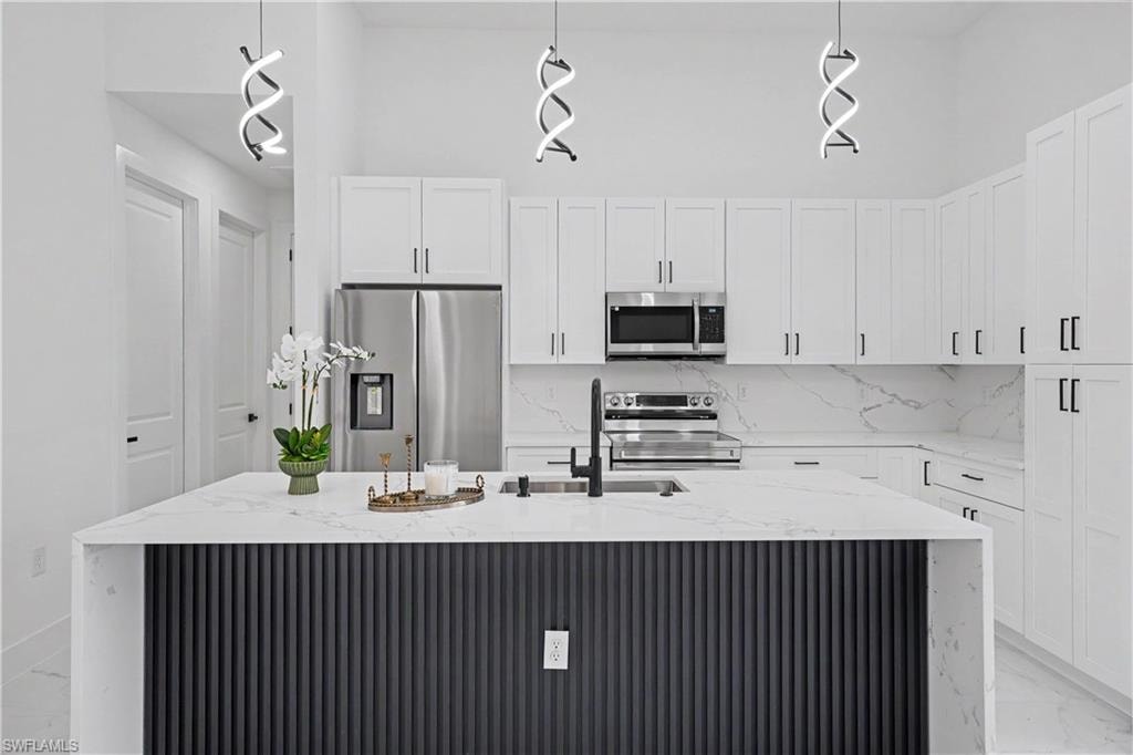 Contemporary kitchen featuring white shaker-style cabinetry, quartz countertops, a waterfall-edge island with vertical ribbed paneling in charcoal finish, stainless steel appliances including a side-by-side refrigerator and built-in microwave, and pendant lighting with modern geometric fixtures suspended above the workspace
