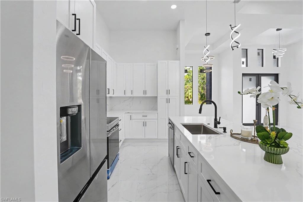 Modern galley kitchen featuring white shaker cabinetry, stainless steel appliances including a French-door refrigerator and built-in oven, white quartz countertops with waterfall edge, matte black faucet with pull-down sprayer, pendant lighting, and light-filled layout with natural light from adjoining windows