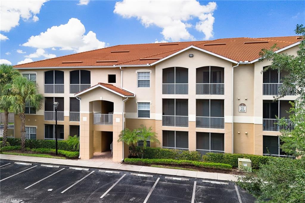 Mediterranean-inspired residential complex featuring cream stucco exterior with terracotta tile roofing, multi-level design with recessed balconies and black-framed windows, manicured landscaping with mature palms and greenery, and dedicated parking area