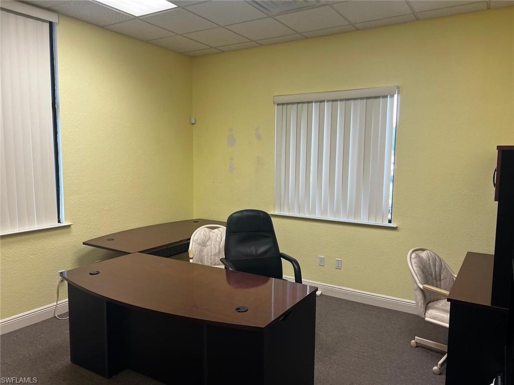 Professional office space featuring a curved executive desk with dark wood-finish top, recessed lighting throughout the ceiling, dual vertical blinds at windows providing light control, neutral yellow walls, and gray commercial-grade flooring