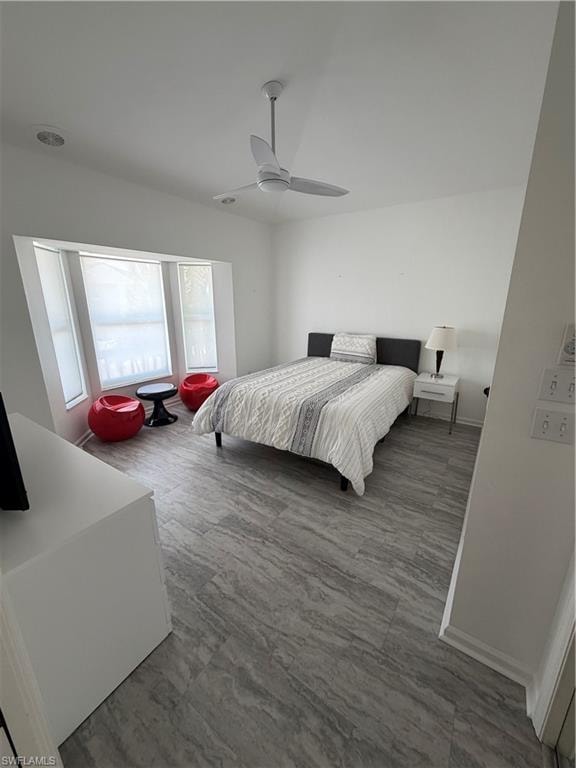 Bright bedroom featuring expansive windows with natural light, wood-finish flooring throughout, ceiling fan, and minimalist design with clean white walls and simple furnishings arrangement