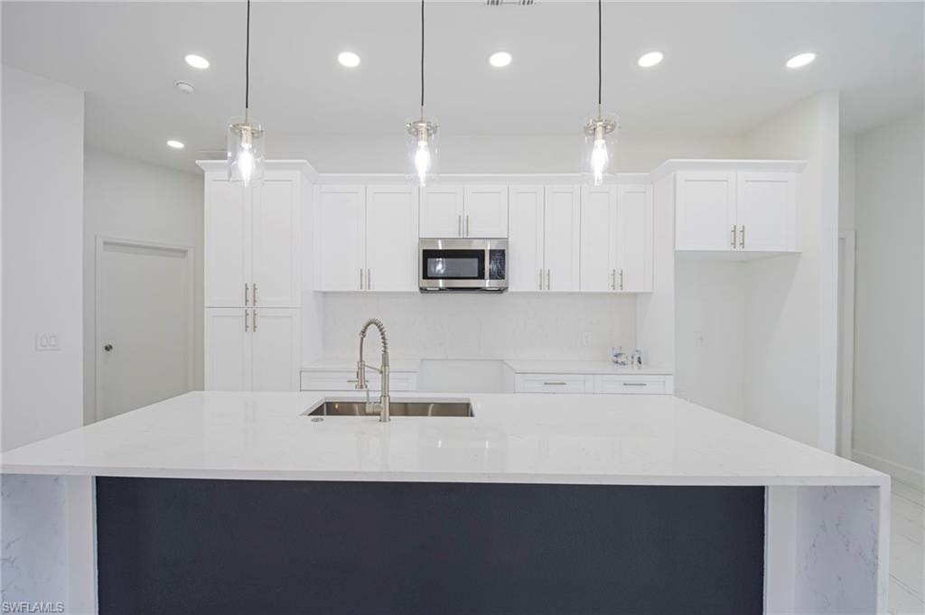 Kitchen with light stone counters, an island with sink, stainless steel microwave, pendant lighting, and two tone cabinets