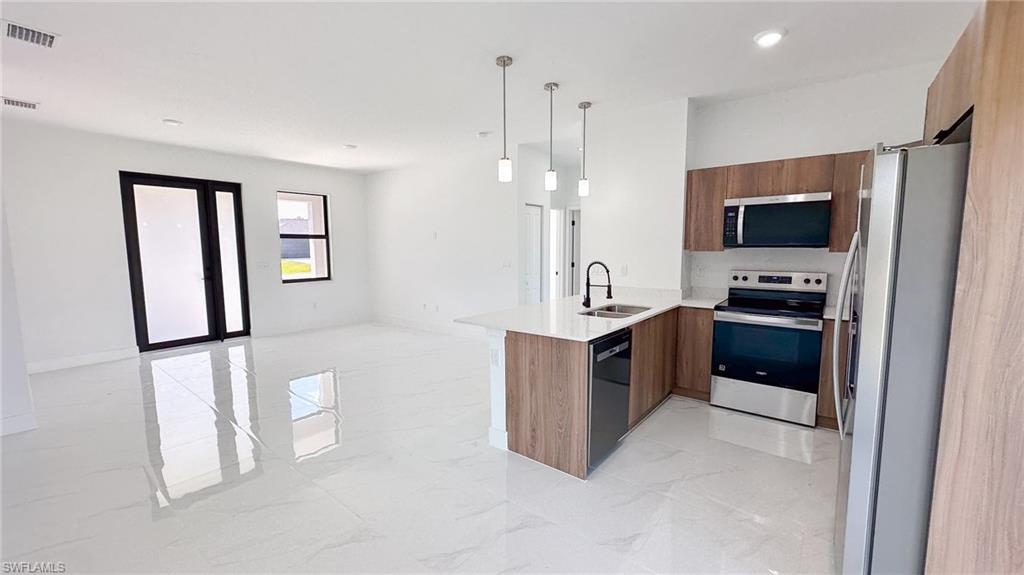 Modern kitchen featuring wood-finish cabinetry, stainless steel appliances including a gas range and refrigerator, a center island with pendant lighting, and polished white flooring throughout the open-concept space