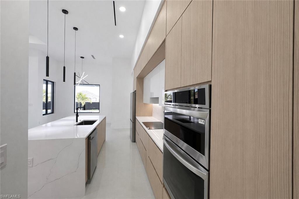 Kitchen with modern cabinets, stainless steel appliances, light wood finish cabinets, light stone counters, and hanging light fixtures