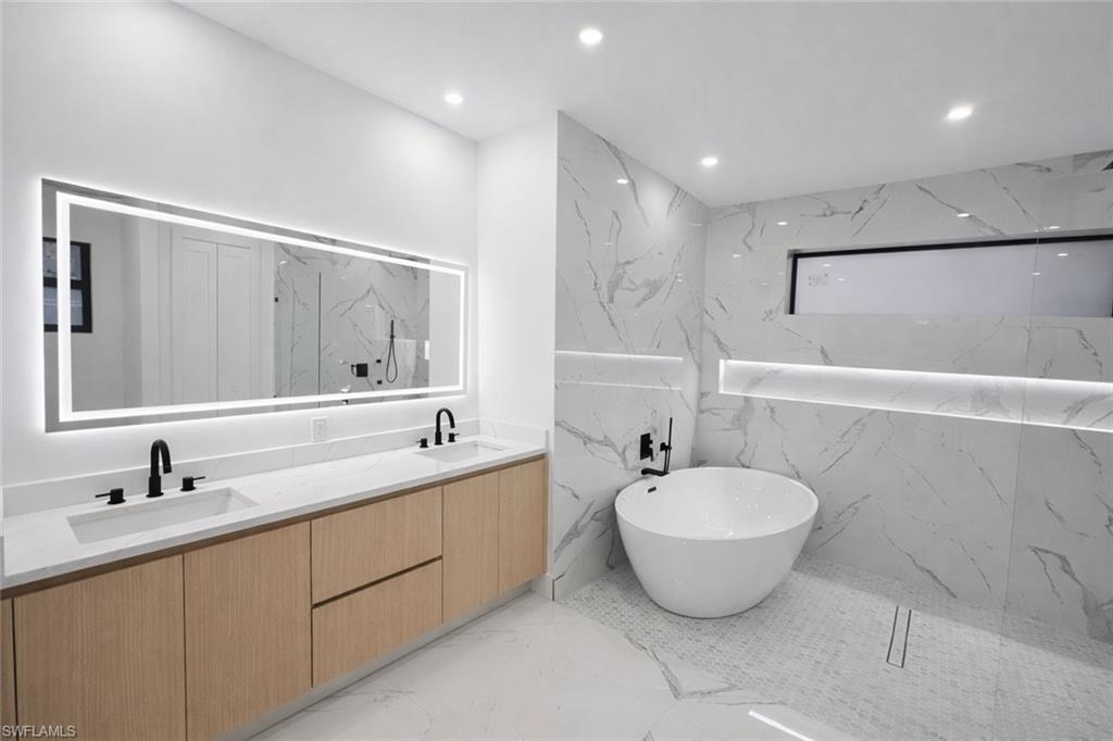Full bath with stone wall, a freestanding bath, double vanity, recessed lighting, and light marble finish floors