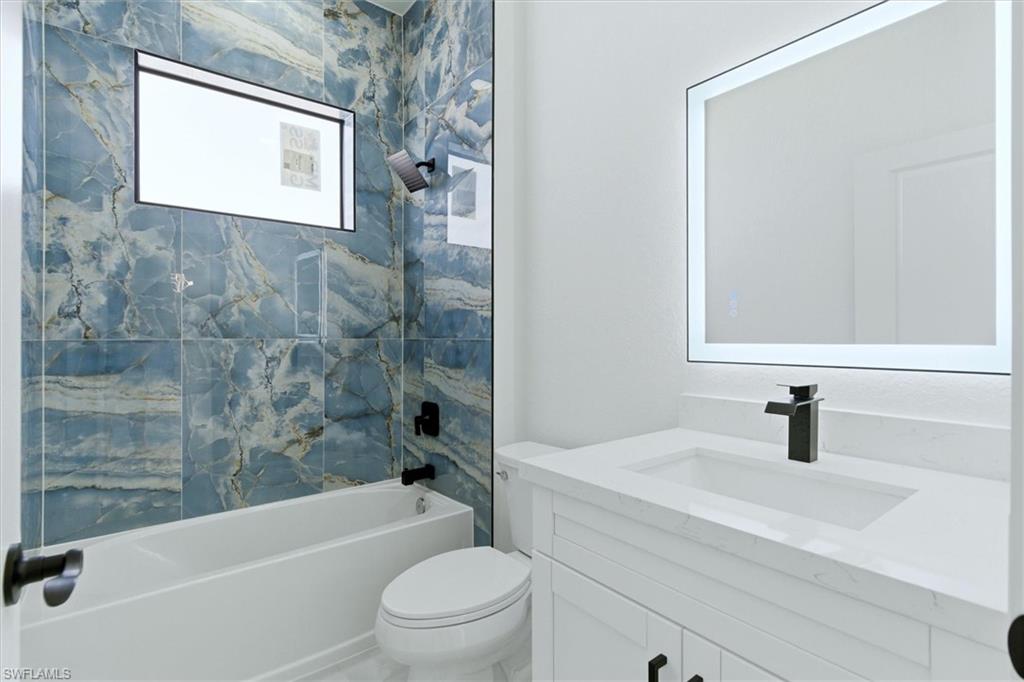 Bathtub with blue veined tile surround, matte black fixtures, built-in window, and white vanity with an integrated sink and illuminated mirror
